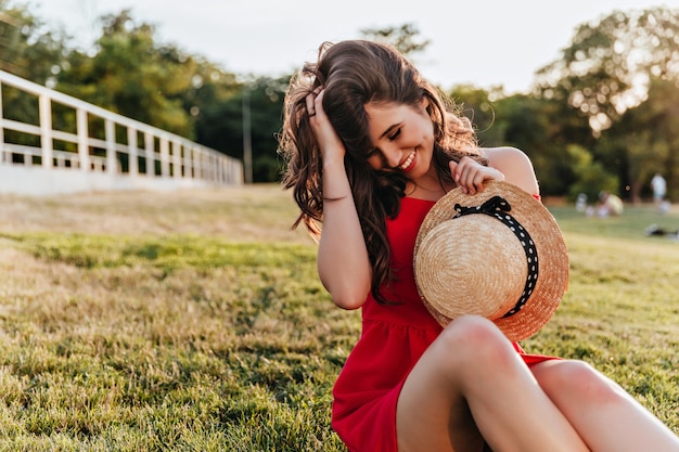 Emocjonalna brunetka dziewczyna w eleganckim kapeluszu ciesząc się wypoczynkiem w parku. Urocza dama w czerwonym stroju siedząca na trawie i dotykająca włosów.
