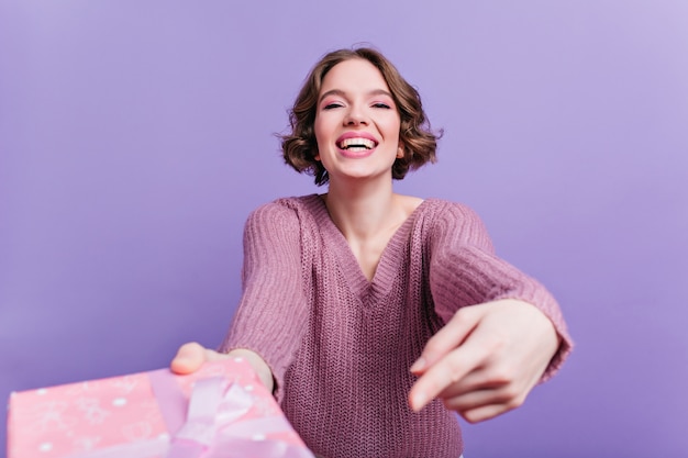 Emocjonalna dziewczyna z ładnym uśmiechem trzymając różowy prezent na fioletowej ścianie. Oszałamiająca krótkowłosa modelka w swetrze z prezentem.