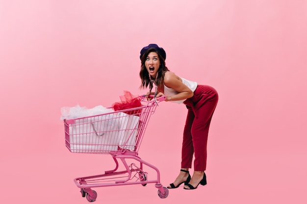 Emocjonalna kobieta w berecie z różowym wózkiem na zakupy. Dziewczyna krzyczy na białym tle stylowy nowoczesny strój.