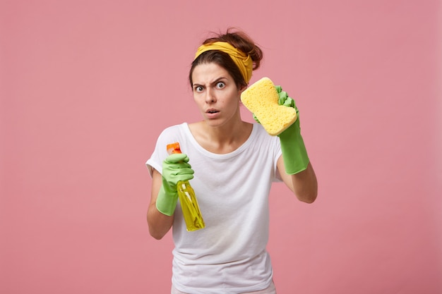 Emocjonalna Urocza Kobieta Pozująca W Zielonych Gumowych Rękawiczkach, Wyposażona W żółtą Gąbkę I Detergent W Sprayu, Gotowa Do Sprzątania I Sprzątania Domu, Patrząc Z Zabawnym Wyrazem Twarzy