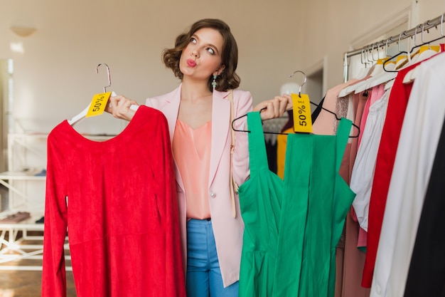 Emocjonalna zabawna atrakcyjna kobieta trzymając kolorowe sukienki na wieszaku w sklepie odzieżowym