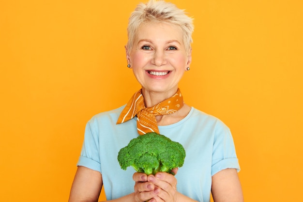Energiczna piękna kobieta w średnim wieku z krótkimi siwymi włosami, odizolowana z zielonymi brokułami w dłoniach, zamierzająca zrobić zdrową, organiczną sałatkę.