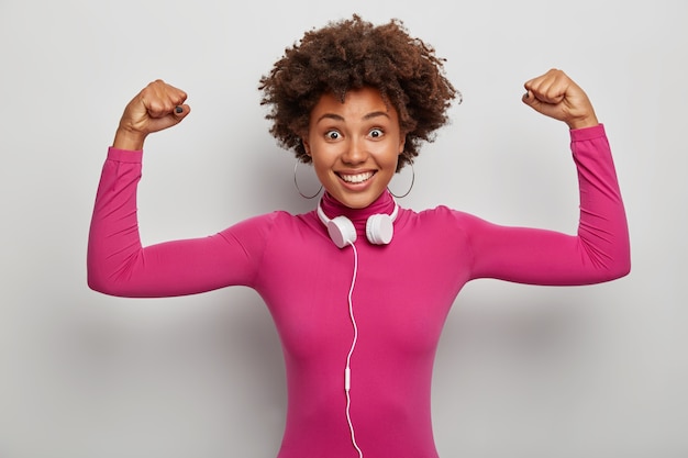 Energiczna, potężna afroamerykańska dama podnosi ramiona, by pokazać mięśnie i siłę, szeroko się uśmiecha, nosi słuchawki stereo na szyi