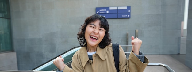 Bezpłatne zdjęcie entuzjastyczna azjatycka dziewczyna stoi na ulicy i robi gest świętowania, mówiąc „tak” triumfując