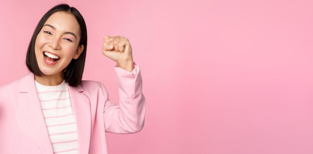 Entuzjastyczna azjatycka kobieta korporacyjna bizneswoman podnosząca rękę w górę i dopingująca triumfującą wygraną i świętującą stojącą na różowym tle