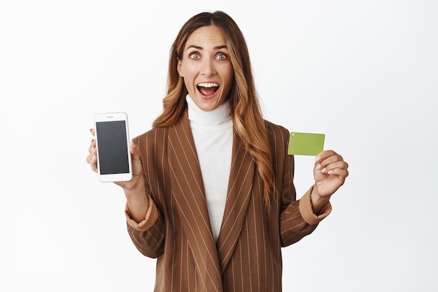 Entuzjastyczna bizneswoman pokazująca interfejs aplikacji na ekranie telefonu i uśmiechając się zdumiony białym tłem karty kredytowej