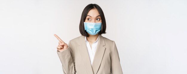 Entuzjastyczna bizneswoman wskazując palcami w lewo, nosząca medyczną maskę na twarz ze stoiska pandemicznego