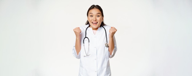 Entuzjastyczna kobieta azjatycka lekarka świętuje patrząc z nadzieją i szczęściem przed kamerą triumfując stojąc w mundurze medycznym na białym tle