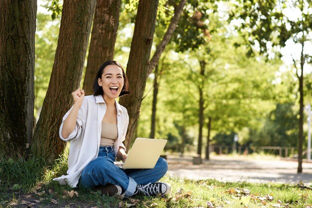 Entuzjastyczna młoda azjatycka dziewczyna siedzi z laptopem obok drzewa w zielonym słonecznym parku świętując triumf