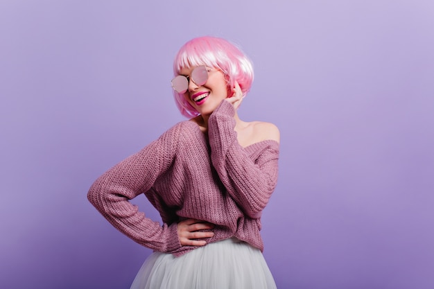 Entuzjastyczna modelka w swetrze i spódnicy tańczy i uśmiecha się. Zainspirowana młoda kobieta z różowymi włosami na fioletowej ścianie.