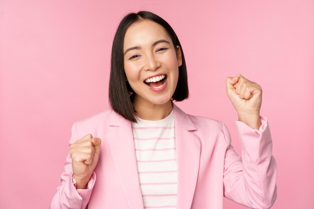 Entuzjastyczna sprzedawczyni azjatycka kobieta korporacyjna mówi tak, osiągaj cel i świętuje triumf patrząc z radością i uśmiechając się stojąc na różowym tle