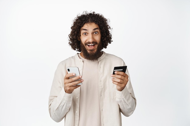Entuzjastyczny arabski facet trzymający telefon komórkowy i kartę kredytową uśmiechający się zdumiony na białym tle aparatu