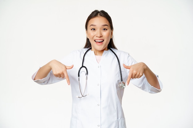 Entuzjastyczny personel medyczny, azjatycka pracownica opieki zdrowotnej, wskazująca palcami w dół i uśmiechnięta zdumiona, pokazująca rabaty, sprzedaż w klinice, białe tło.