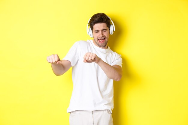 Fajny facet słuchanie muzyki w słuchawkach i taniec, stojąc w białych ubraniach na żółtym tle studia.