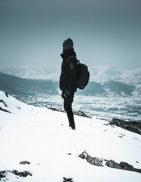 Fajny turysta w masce i ciemnych ubraniach stojący samotnie na zaśnieżonym stromym wzgórzu w górach