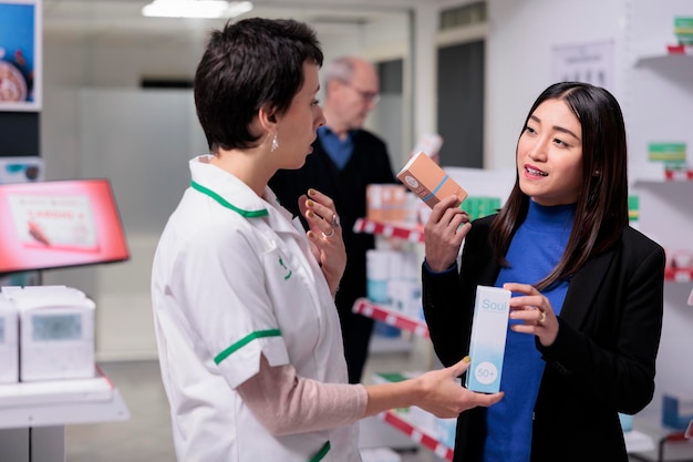 Farmaceuta Polecający Krem Z Filtrem Przeciwsłonecznym Młodej Azjatyckiej Kobiecie W Aptece. Klient Trzymający Pakiety Produktów Do Pielęgnacji Skóry, Wybierający Między Dwoma Balsamami Do Opalania I Konsultujący Się Z Asystentem Apteki