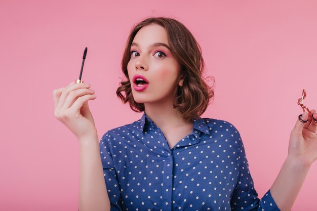 Fascynująca młoda kobieta w eleganckiej bluzce pozuje podczas robienia makijażu. Ekstatyczna zaskoczona dziewczyna trzyma pędzel do tuszu do rzęs na różowej ścianie.