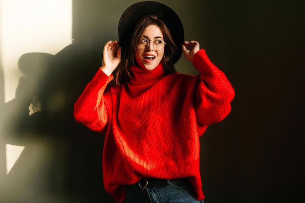 Fashionmonger w kapeluszu z szerokim rondem i okularach pozuje w studio na tle własnego cienia Dziewczyna w czerwonym swetrze oversize wygląda na bok