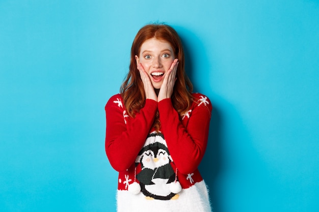 Ferie zimowe i koncepcja Wigilię Bożego Narodzenia. Podekscytowana ruda dziewczyna w xmas sweter patrząc zaskoczony na aparat, stojąc na niebieskim tle.