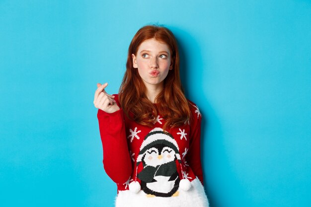 Ferie zimowe i koncepcja Wigilię Bożego Narodzenia. Urocza rudowłosa kobieta w świątecznym swetrze, pokazująca znak serca i myśląca, patrząca na logo w lewym górnym rogu, niebieskie tło