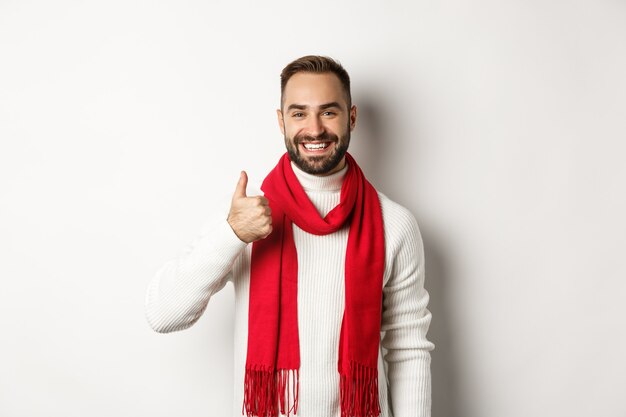 Ferie zimowe i koncepcja zakupów. Pewnie przystojny mężczyzna pokazując kciuk do góry, stojący w świąteczny sweter i czerwony szalik, białe tło.