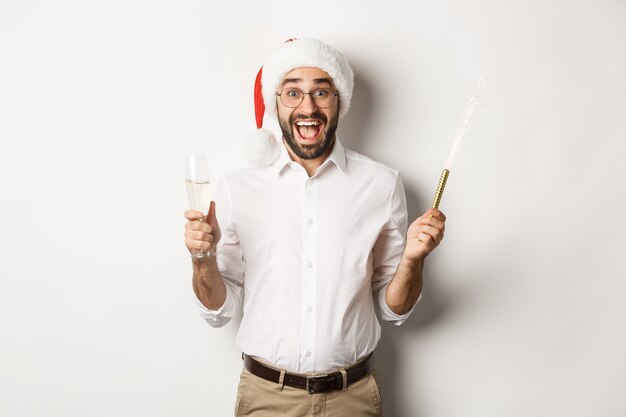 Ferie zimowe i uroczystości. Szczęśliwy facet w czapce Mikołaja radujący się na imprezie noworocznej, pijący szampana i krzyczący z radości