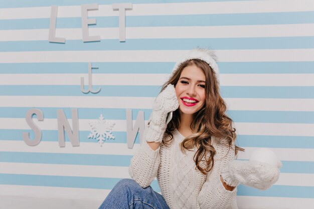 Figlarna, fajna modelka z uśmiechem, patrząca, trzymająca zwitek śniegu w ciepłych rękawiczkach. Portret młodej dziewczyny w pokoju z pasiastymi ścianami