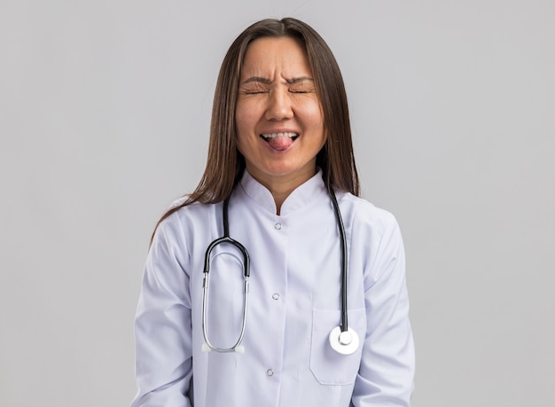 Figlarna młoda azjatycka lekarka nosząca medyczną szatę i stetoskop pokazujący język z zamkniętymi oczami na białym tle na białej ścianie