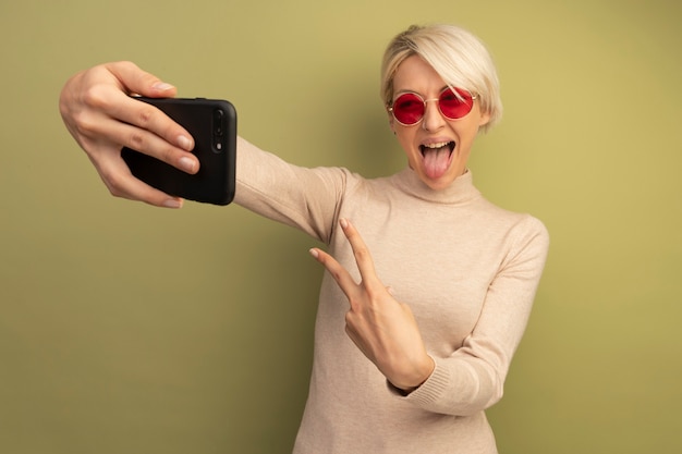 Figlarna młoda blondynka w okularach przeciwsłonecznych robi znak pokoju pokazujący język biorący selfie na oliwkowozielonej ścianie