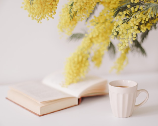 Filiżanka kawy i książka na białym biurku z mimozy bukiet żółte kwiaty.