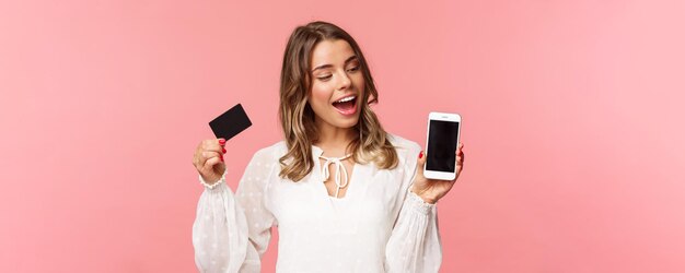 Finanse koncepcja zakupów i technologii Zbliżenie portret podekscytowanej ładnie wyglądającej blond dziewczyny w białej sukni odnawia jej wiosenną garderobę z kupowaniem ubrań online za pomocą smartfona, trzymaj kartę kredytową