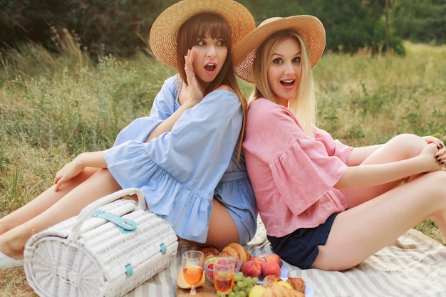 Bezpłatne zdjęcie fotografia mody plenerowej dwóch atrakcyjnych kobiet w słomkowym kapeluszu i letnich ubraniach, które zachwycają piknika.