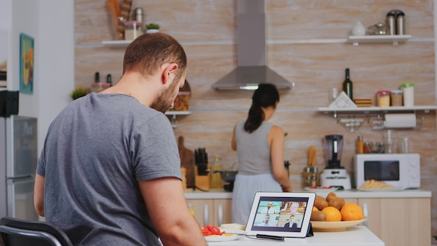 Freelancer podczas wideokonferencji na komputerze typu tablet, podczas gdy żona gotuje śniadanie w kuchni. Przedsiębiorca przy filiżance kawy podczas wideokonferencji ze współpracownikami.