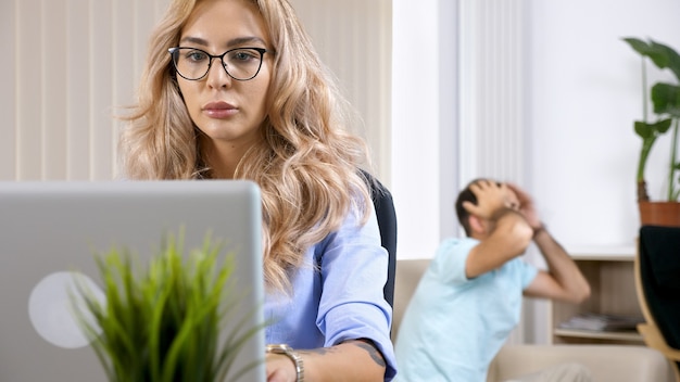 Freelancerka pracująca na laptopie w domu, podczas gdy mąż w tle ogląda telewizję