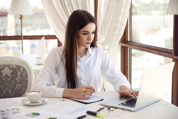 Frontowy widok młody brunetka bizneswoman który pracuje na laptopie i pisze coś puszkowi