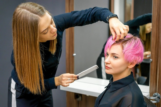 Fryzjer kobiece stylizacja krótkie różowe włosy młodej białej kobiety z rąk i grzebienia w salonie fryzjerskim.