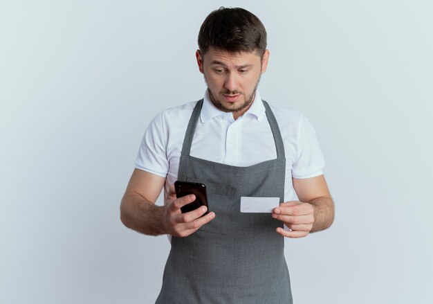 Fryzjer mężczyzna w fartuch loking mylić trzymając smartfon i stojącą kartę kredytową na białym tle