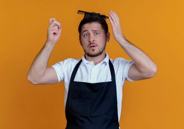 Fryzjer mężczyzna w fartuch ze szczotką do włosów we włosach patrząc na kamery zdezorientowany stojąc na pomarańczowym tle