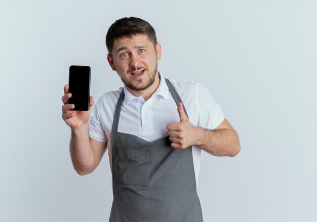 fryzjer mężczyzna w fartuchu pokazuje smartfon pokazując kciuki do góry uśmiechnięty pewny siebie stojący nad białą ścianą