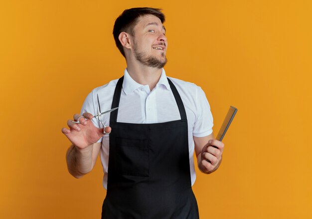 fryzjer mężczyzna w fartuchu trzymając nożyczki i grzebień uśmiechnięty wesoło stojąc nad pomarańczową ścianą