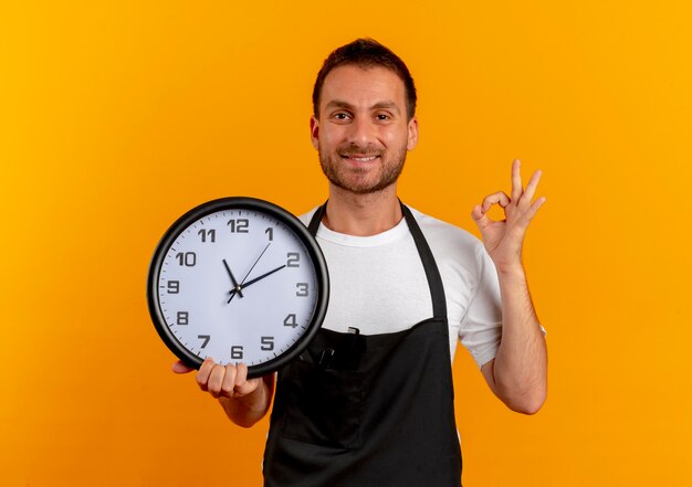 Fryzjer w fartuchu trzyma zegar ścienny patrząc do przodu, uśmiechając się wesoło, pokazując znak ok stojącego nad pomarańczową ścianą