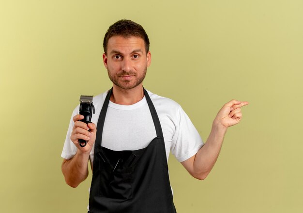 Fryzjer w fartuchu trzymający trymer wskazujący palcem wskazującym w bok, wyglądający pewnie na stojącego nad jasną ścianą
