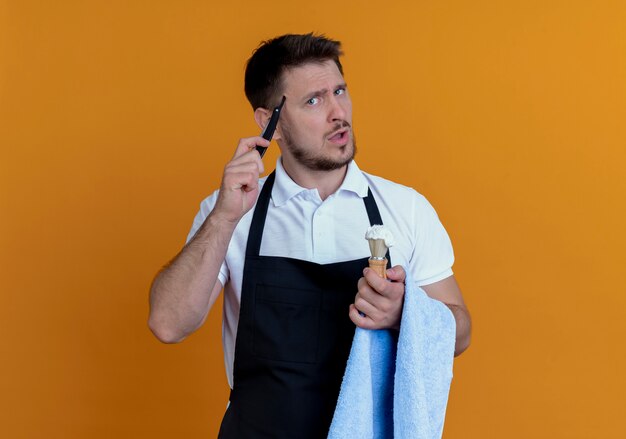 Fryzjer w fartuchu z ręcznikiem na dłoni trzymający pędzel do golenia z pianką i brzytwą patrząc na aparat zdziwiony stojąc na pomarańczowym tle