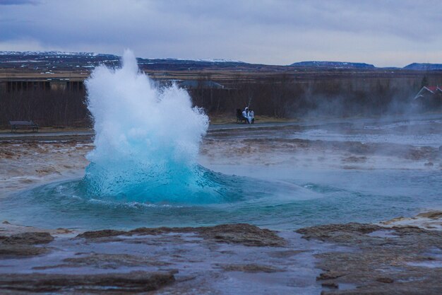 Gejzer Strokkur otoczony wzgórzami pod zachmurzonym niebem wieczorem na Islandii