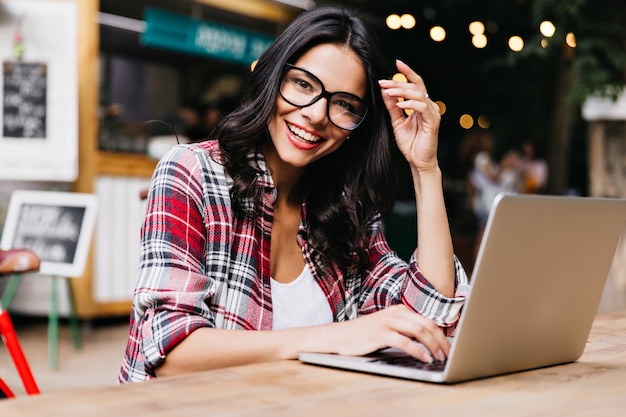 Glamorous kobieta freelancer ciesząc się rano i pracując z laptopem. Zdjęcie wesołej pani Łacińskiej w kraciastej koszuli pozuje w okularach.