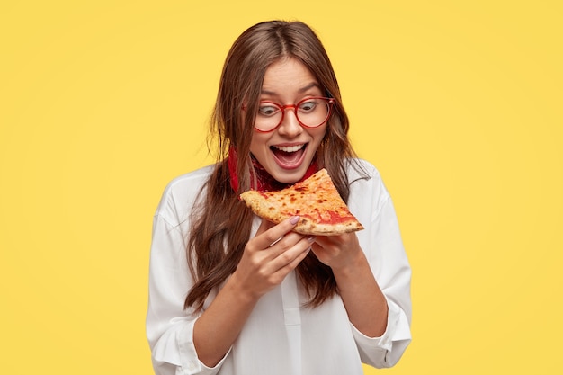 Głodny student szeroko otwiera usta, widząc kawałek pysznej pizzy, chce zjeść, ubrany w białą koszulę, modelki przy żółtej ścianie. Pozytywna kobieta z fast foodem. Ludzie i jedzenie