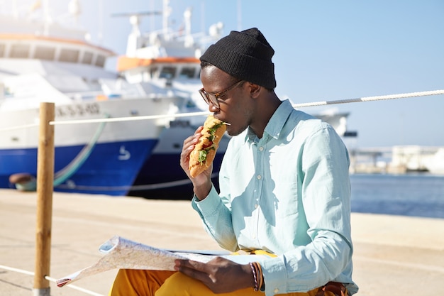 Głodny Turysta Mężczyzna W Modnych Ubraniach I Dodatkach Jedzący Kanapkę