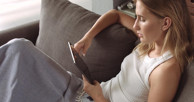 Bezpłatne zdjęcie gorąca blond modelka czyta na swoim tablecie siedząc na ciemnoszarej kanapie pokrytej miękkim narzutem