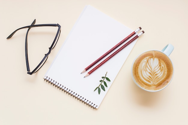 Gorąca kawa cappuccino latte art; okulary i ołówki na białym spiralnym notatniku
