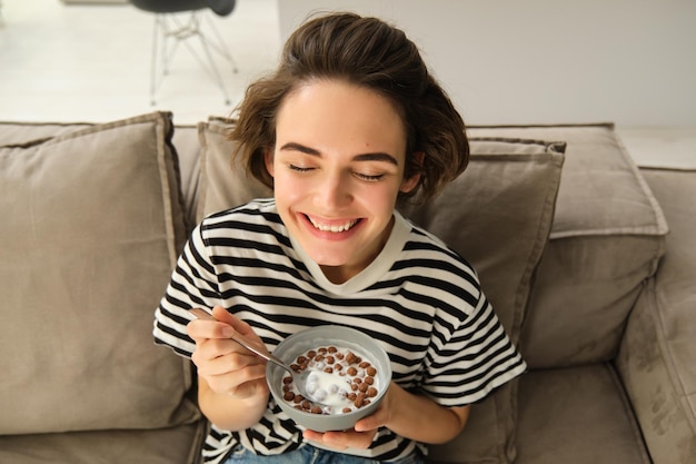 Bezpłatne zdjęcie górny kąt zdjęcia szczęśliwej słodkiej młodej kobiety na kanapie jedzącej miskę płatków zbożowych z mlekiem i uśmiechającej się
